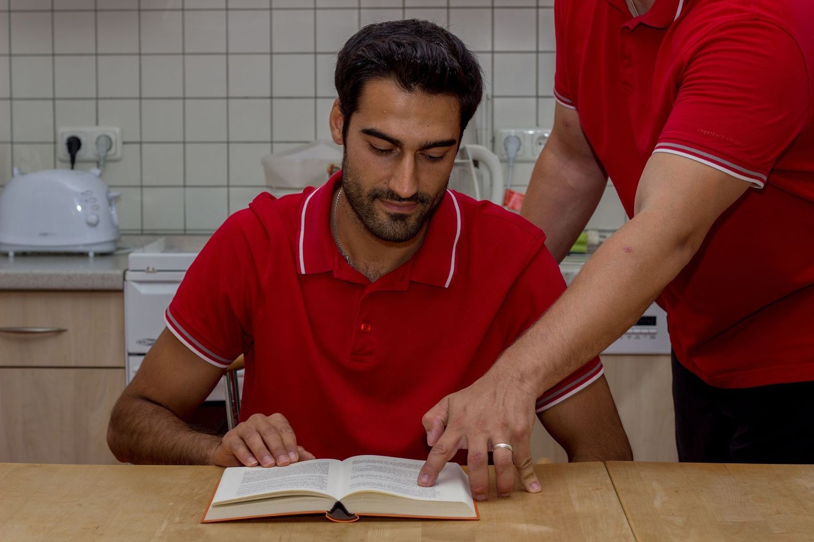 Two men in red shirts engaged in reading and discussion at a kitchen table, focusing on a book.